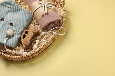 Different baby accessories and clothes in wicker basket on yellow background, above view