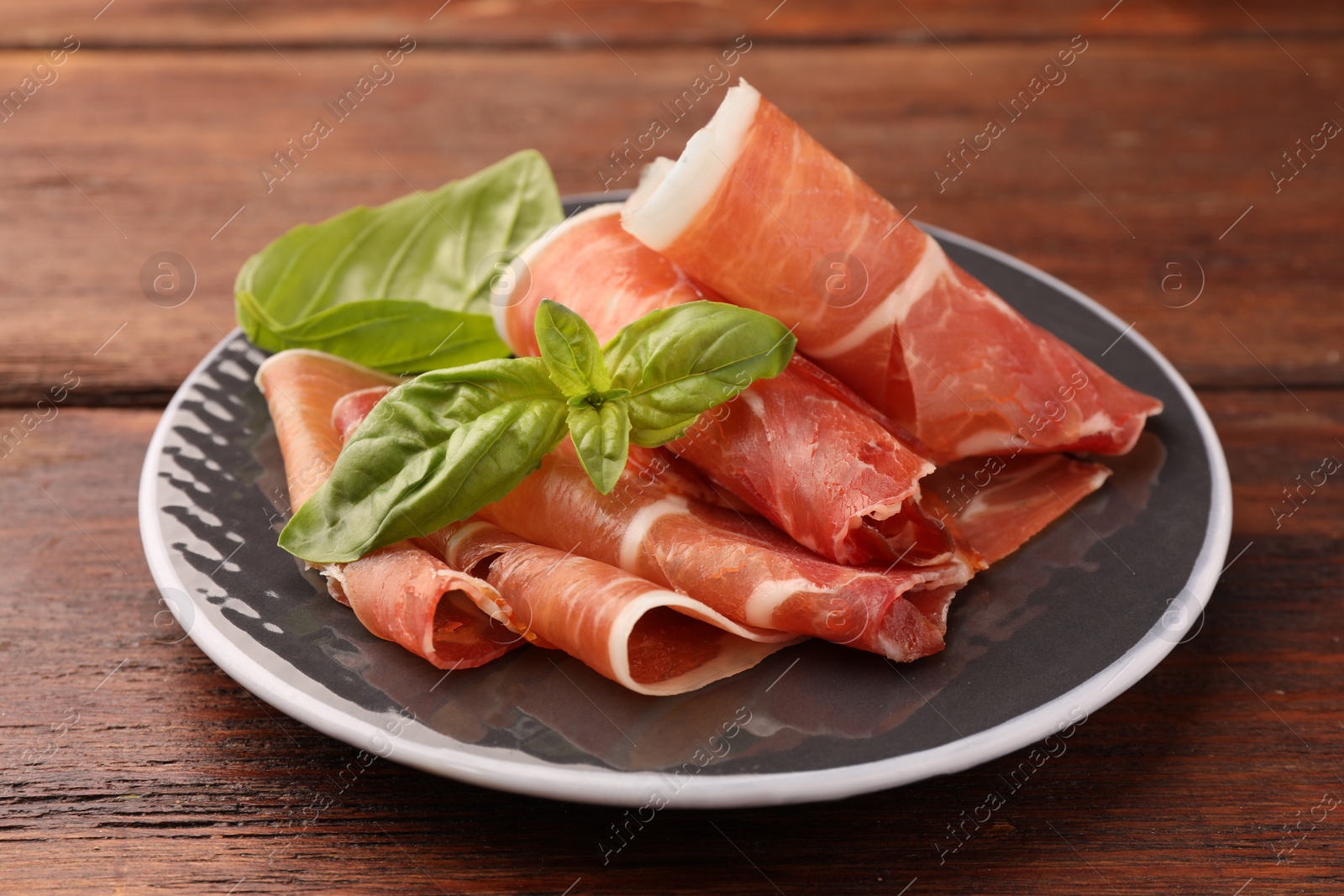 Photo of Plate with rolled slices of delicious jamon and basil on wooden table, closeup