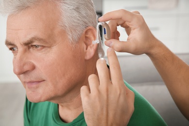 Photo of Young man putting hearing aid in father's ear indoors, closeup