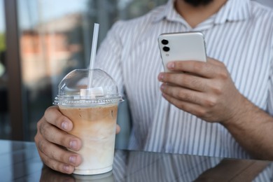 Photo of Man with smartphone and plastic takeaway cup of delicious iced coffee at table in outdoor cafe, closeup