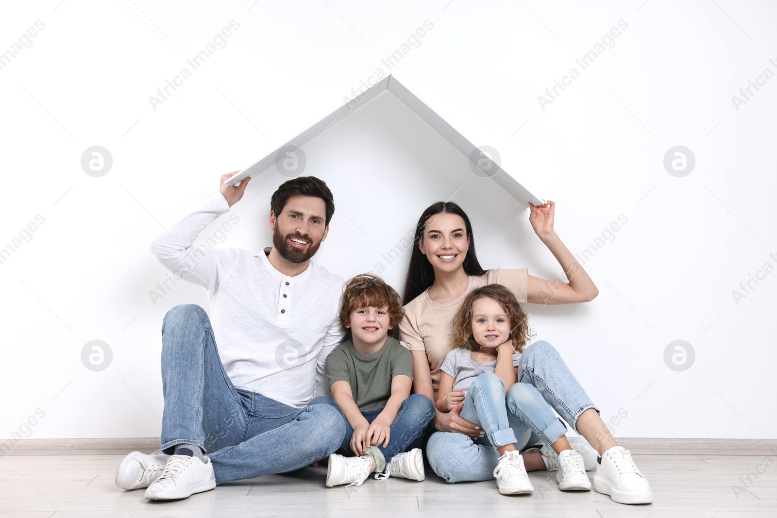 Photo of Family housing concept. Happy woman and her husband holding plastic roof while sitting with kids on floor at home