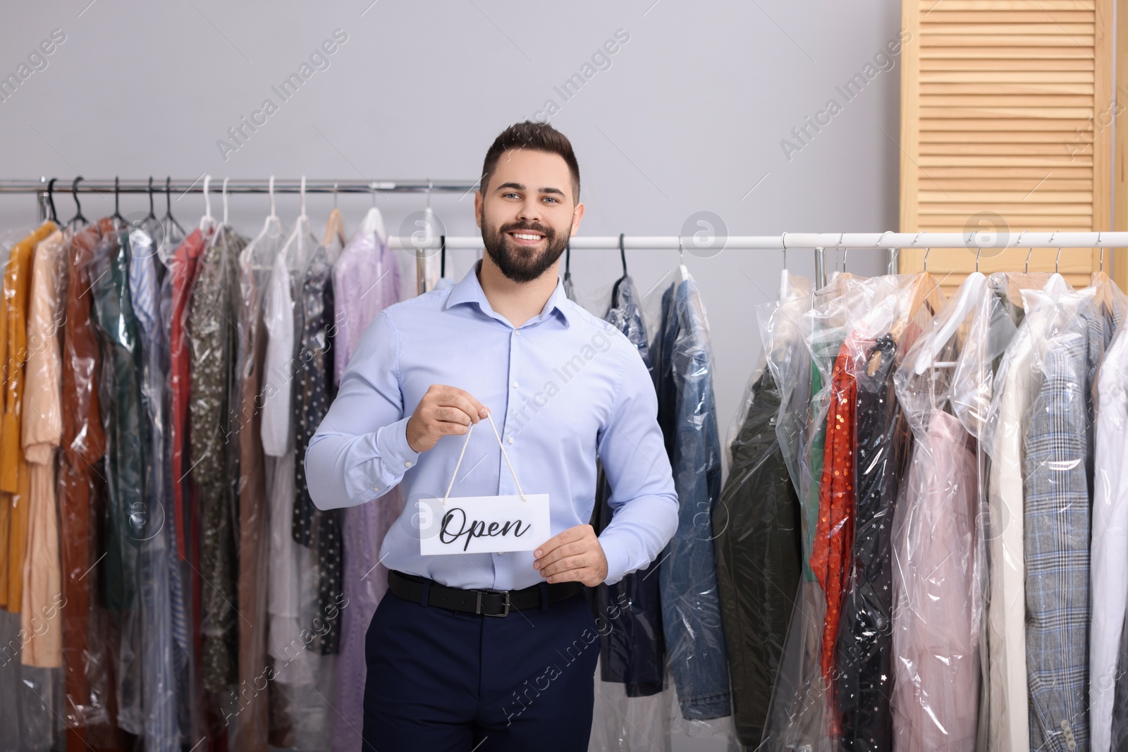 Photo of Dry-cleaning service. Happy worker holding Open sign near racks with clothes indoors