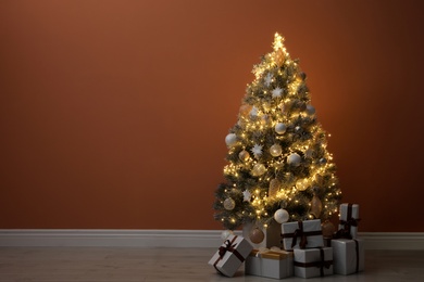 Beautiful decorated Christmas tree with glowing fairy lights and presents near orange wall indoors. Space for text