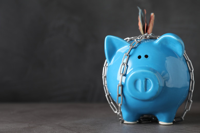 Photo of Piggy bank with steel chain and banknotes on grey stone table, space for text. Money safety concept