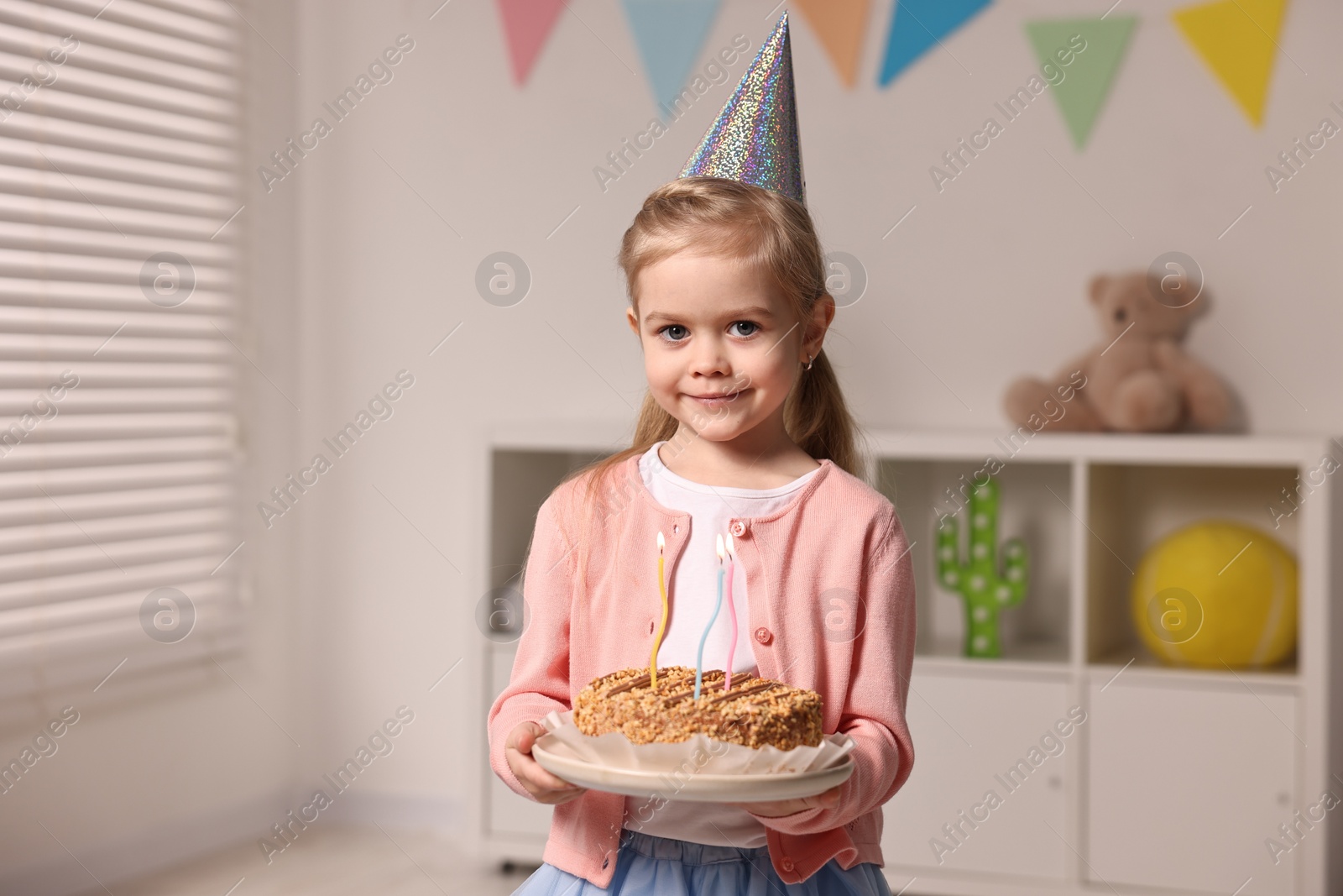Photo of Birthday celebration. Cute little girl in party hat holding tasty cake with burning candles at home