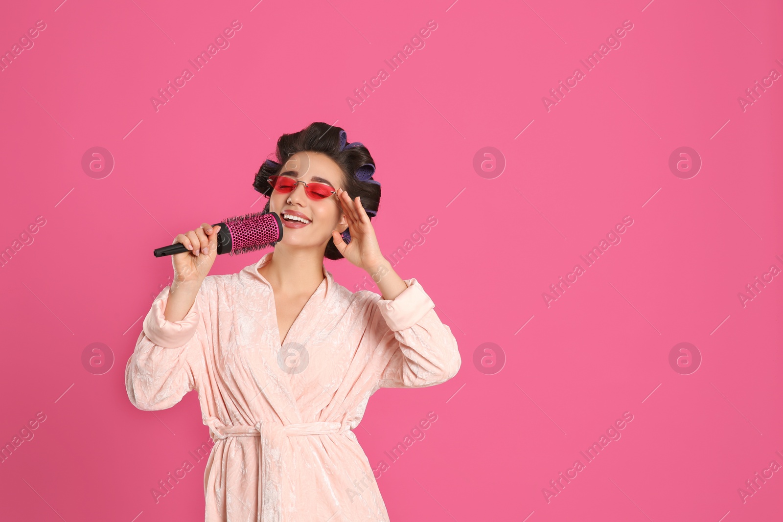 Photo of Happy young woman in bathrobe with hair curlers and sunglasses singing into hairbrush on pink background, space for text