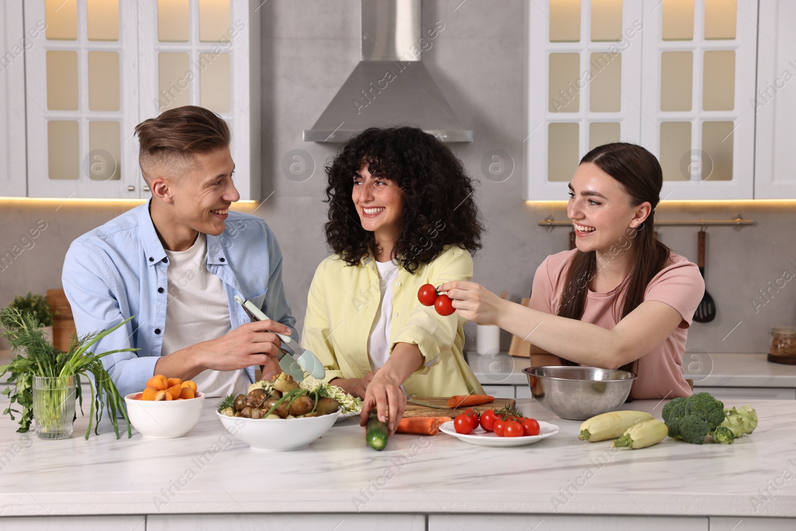 Photo of Friends cooking healthy vegetarian meal at white marble table in kitchen