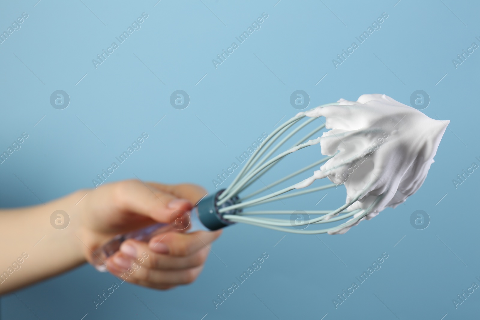 Photo of Woman holding whisk with whipped cream on light blue background, closeup