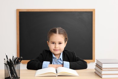 Little school child sitting at desk with books near chalkboard in classroom