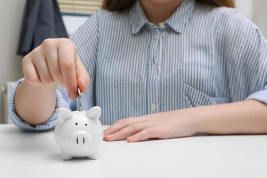 Photo of Woman putting coin into ceramic piggy bank at white wooden table, closeup. Financial savings