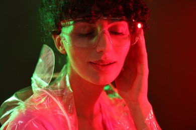 Photo of Beautiful young woman in transparent coat and sunglasses posing on color background in neon lights, closeup