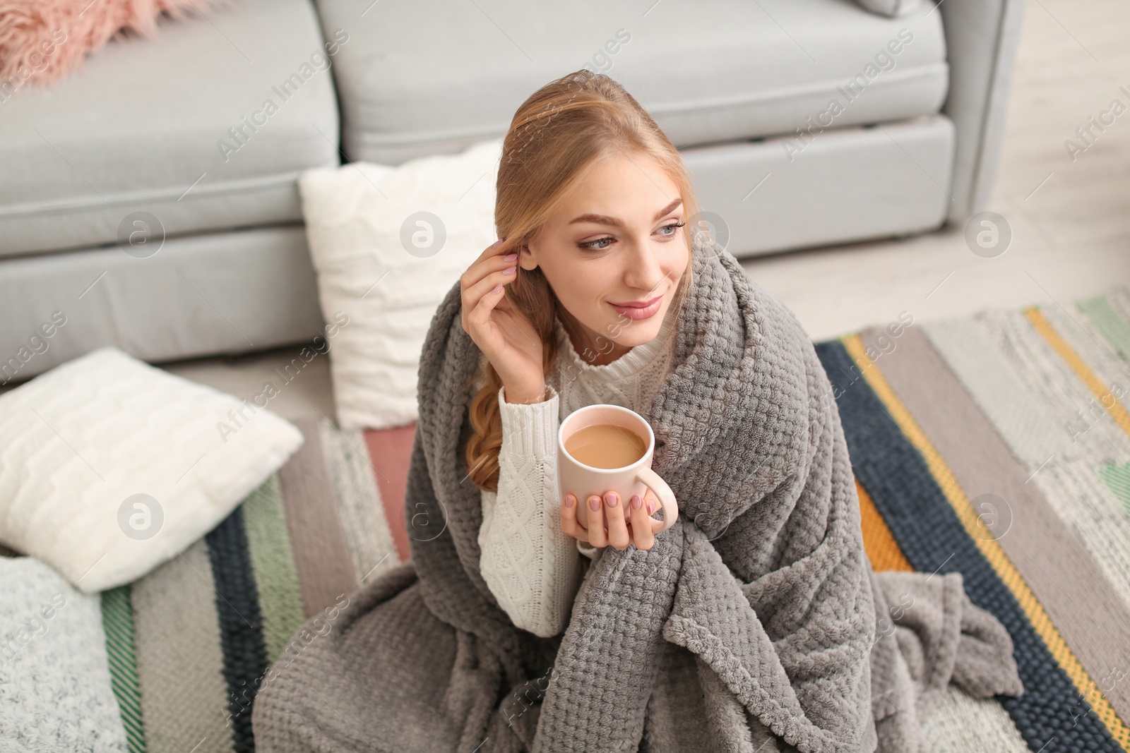 Photo of Beautiful young woman wrapped in plaid sitting with cup of coffee on floor at home. Winter atmosphere