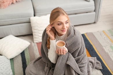 Beautiful young woman wrapped in plaid sitting with cup of coffee on floor at home. Winter atmosphere