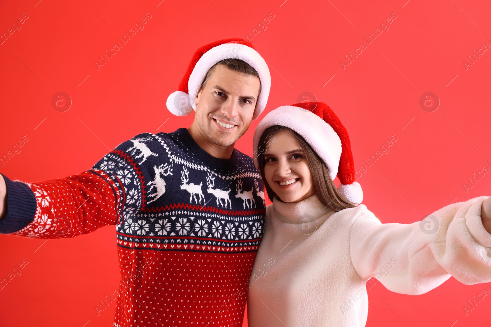 Photo of Beautiful happy couple in Santa hats and Christmas sweaters taking selfie on red background