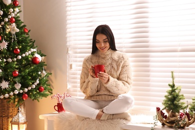 Photo of Young woman with cup of hot drink near Christmas tree at home