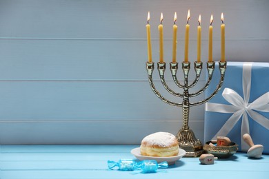 Photo of Hanukkah celebration. Menorah with burning candles, dreidels, donut and gift box on light blue wooden table, space for text