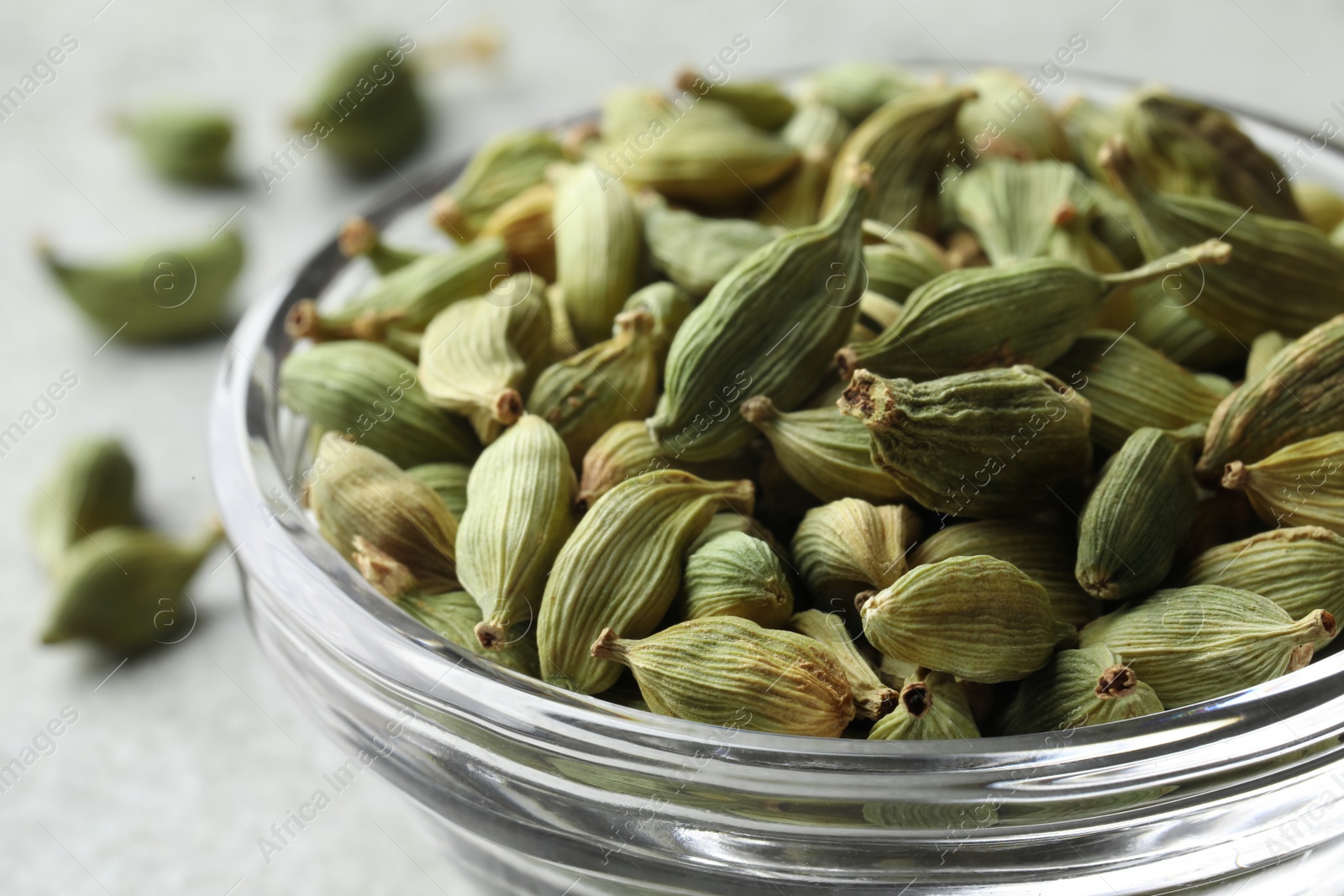 Photo of Glass bowl of dry cardamom pods on light grey table, closeup