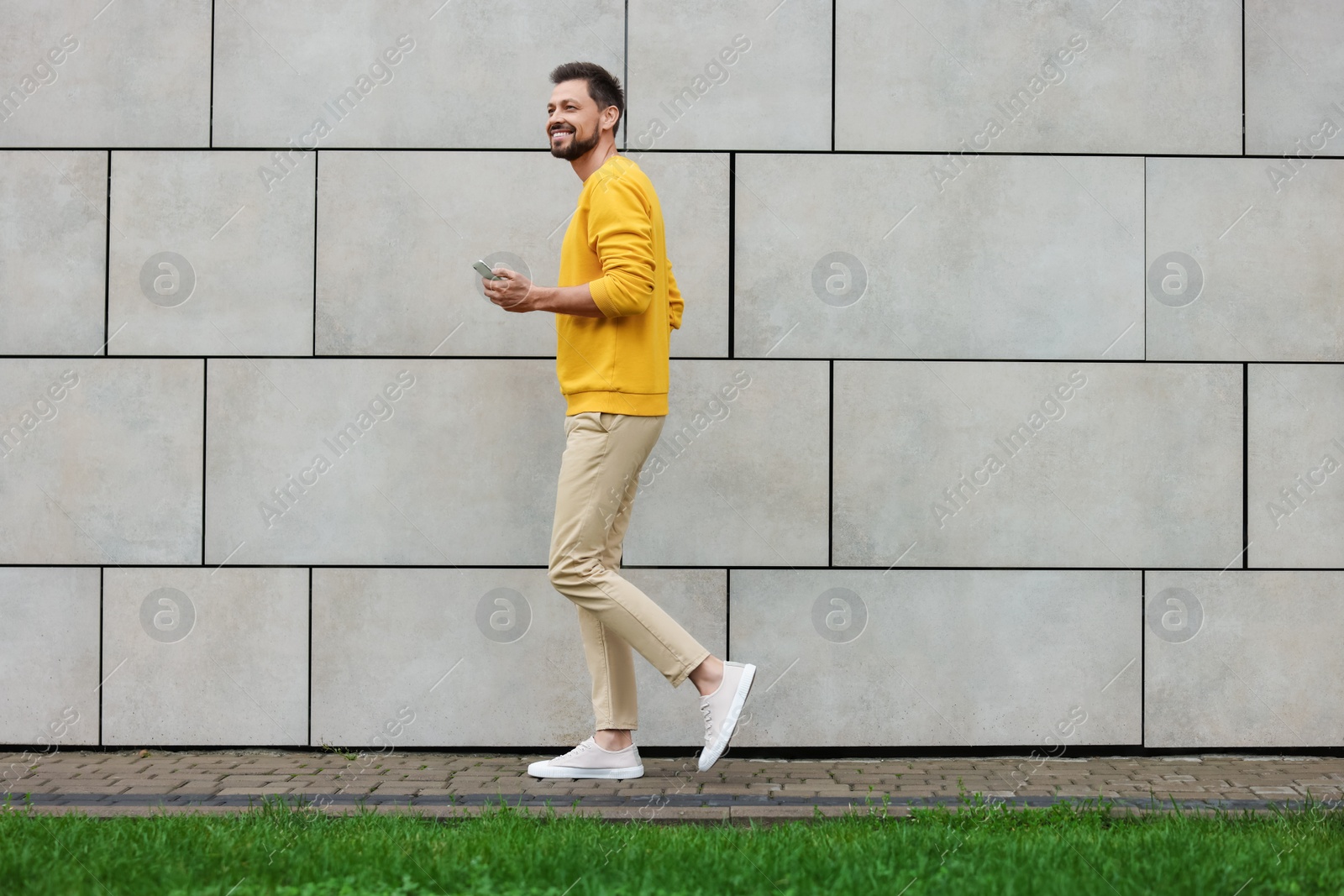 Photo of Handsome man with smartphone walking near grey stone wall outdoors, space for text