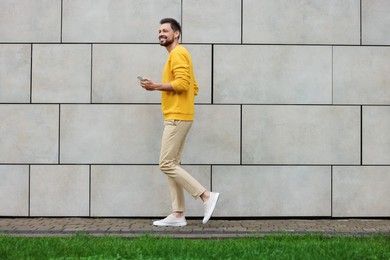Photo of Handsome man with smartphone walking near grey stone wall outdoors, space for text