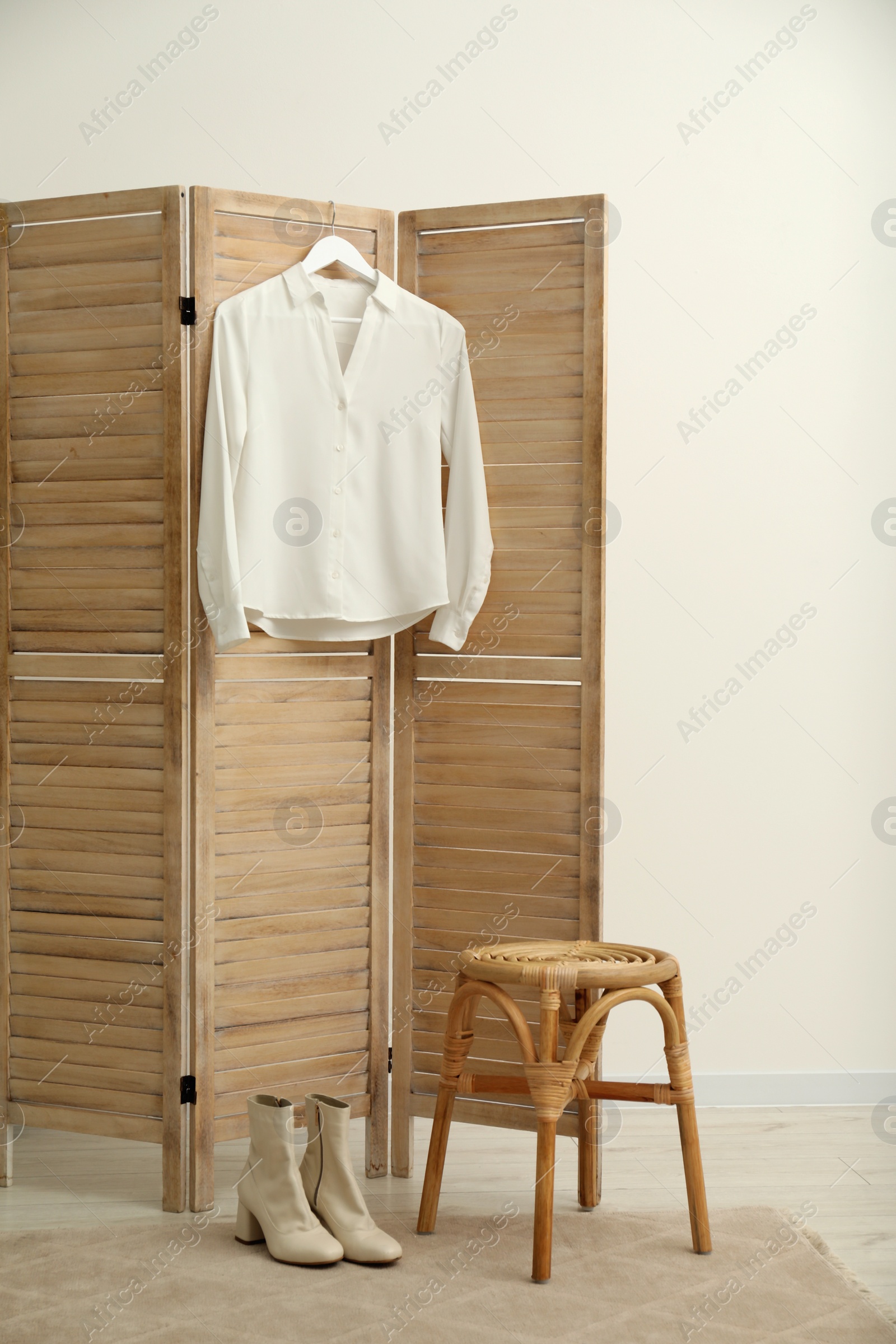 Photo of Stylish white shirt hanging on wooden folding screen, stool and boots indoors