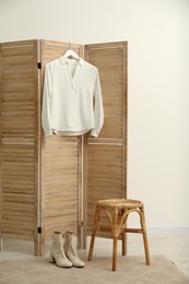 Photo of Stylish white shirt hanging on wooden folding screen, stool and boots indoors