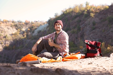 Photo of Male camper with thermos sitting on sleeping bag in wilderness