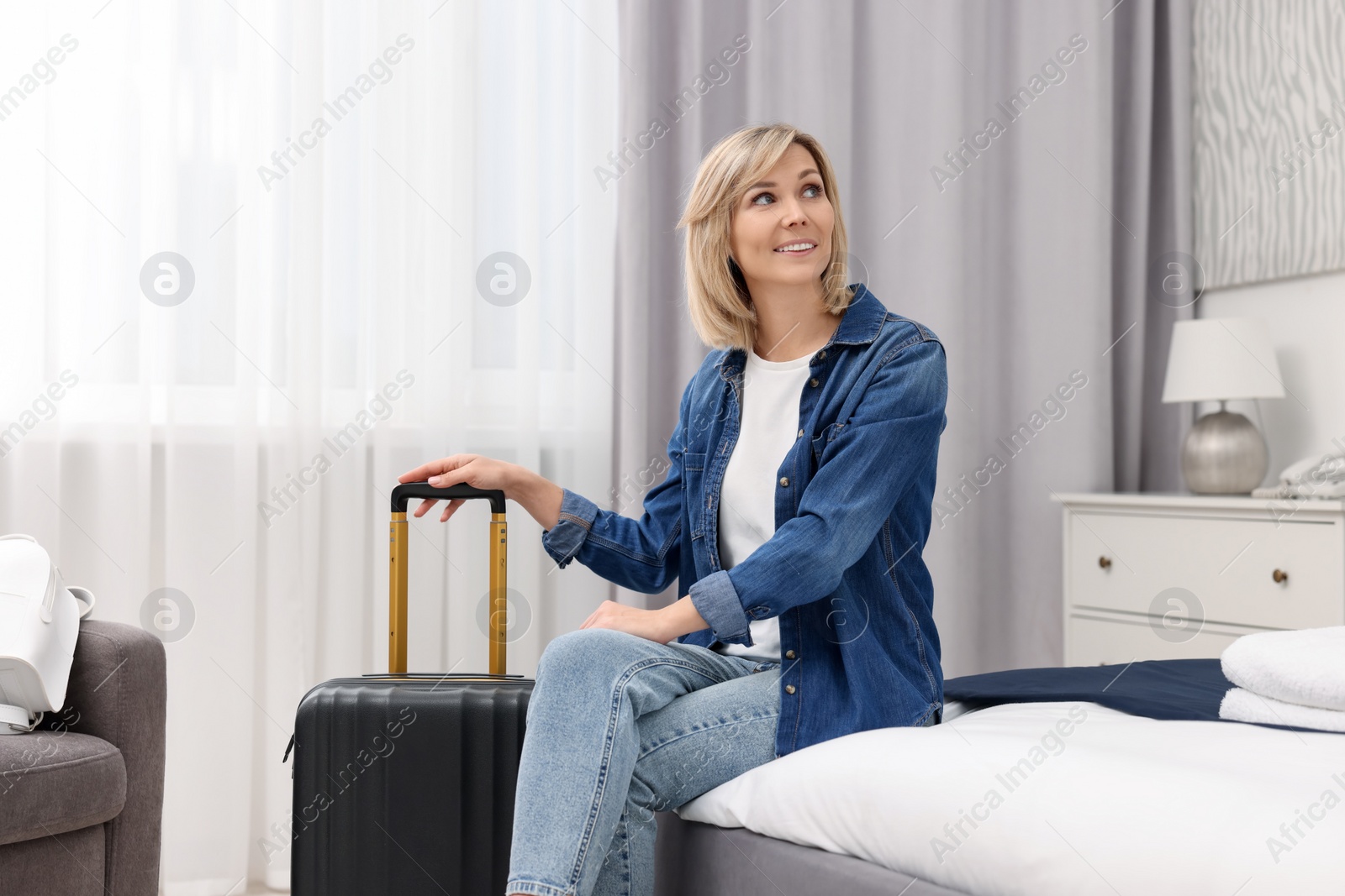 Photo of Smiling guest with suitcase relaxing on bed in stylish hotel room