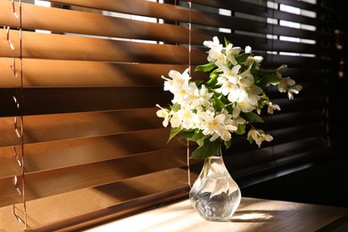 Photo of Bouquet of beautiful jasmine flowers in glass vase on wooden table near window indoors, space for text