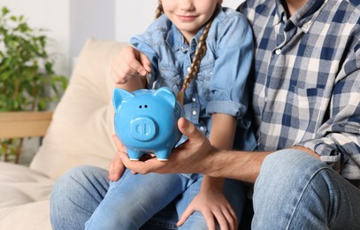 Photo of Little girl with her father putting coin into piggy bank at home, closeup
