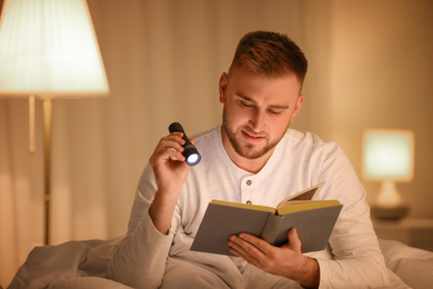 Young man with flashlight reading book in bedroom