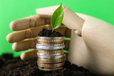 Stack of coins, green plant and wooden mannequin hand on soil against blurred background. Profit concept