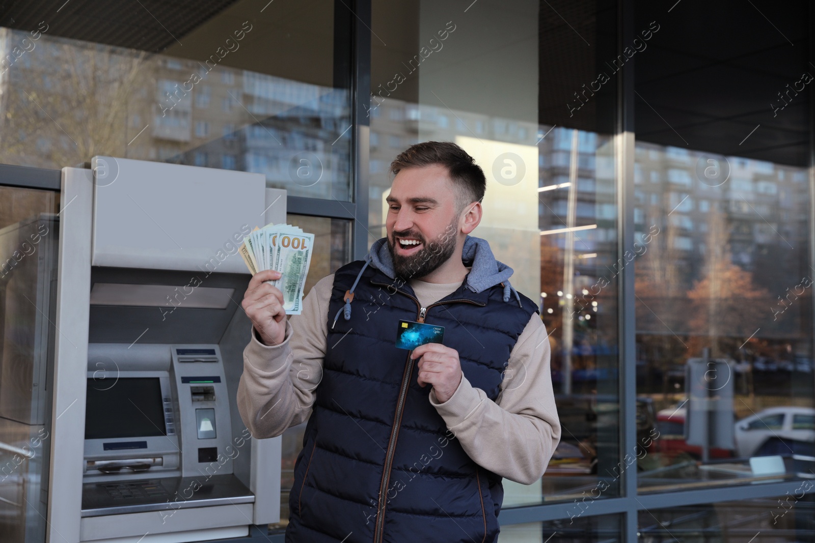 Photo of Excited man with credit card and money near cash machine outdoors