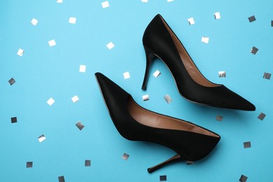 Pair of elegant black high heel shoes and confetti on light blue background, flat lay. Space for text