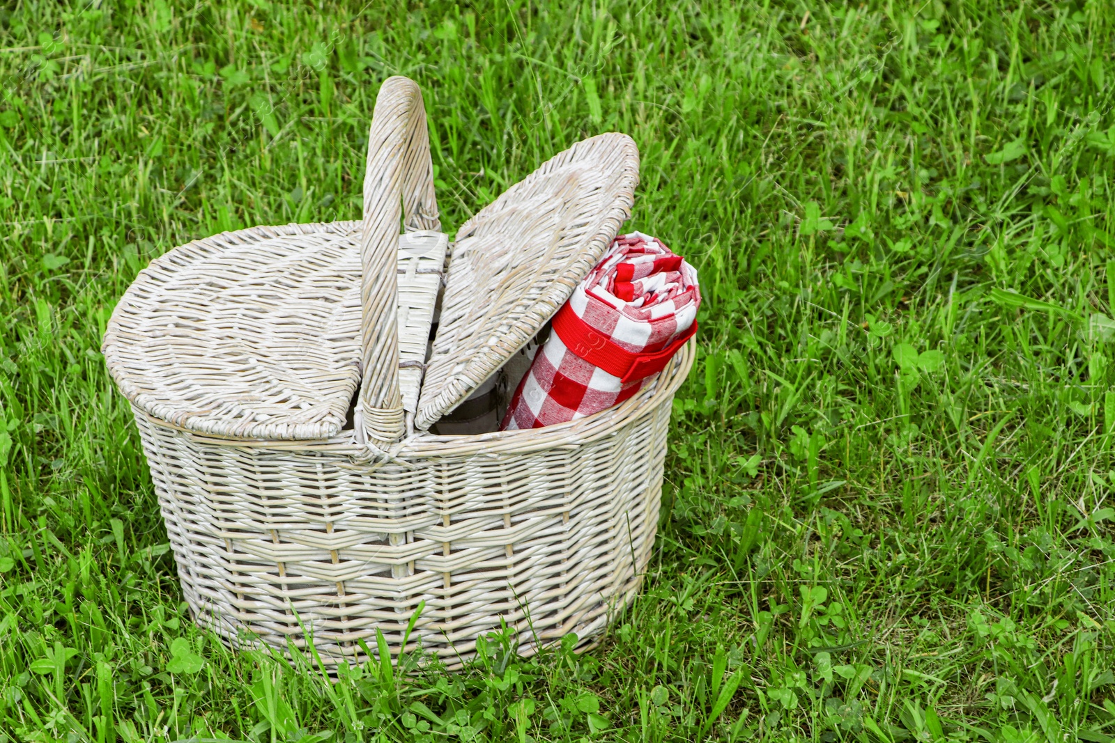 Photo of Rolled checkered tablecloth in picnic basket on green grass outdoors, space for text