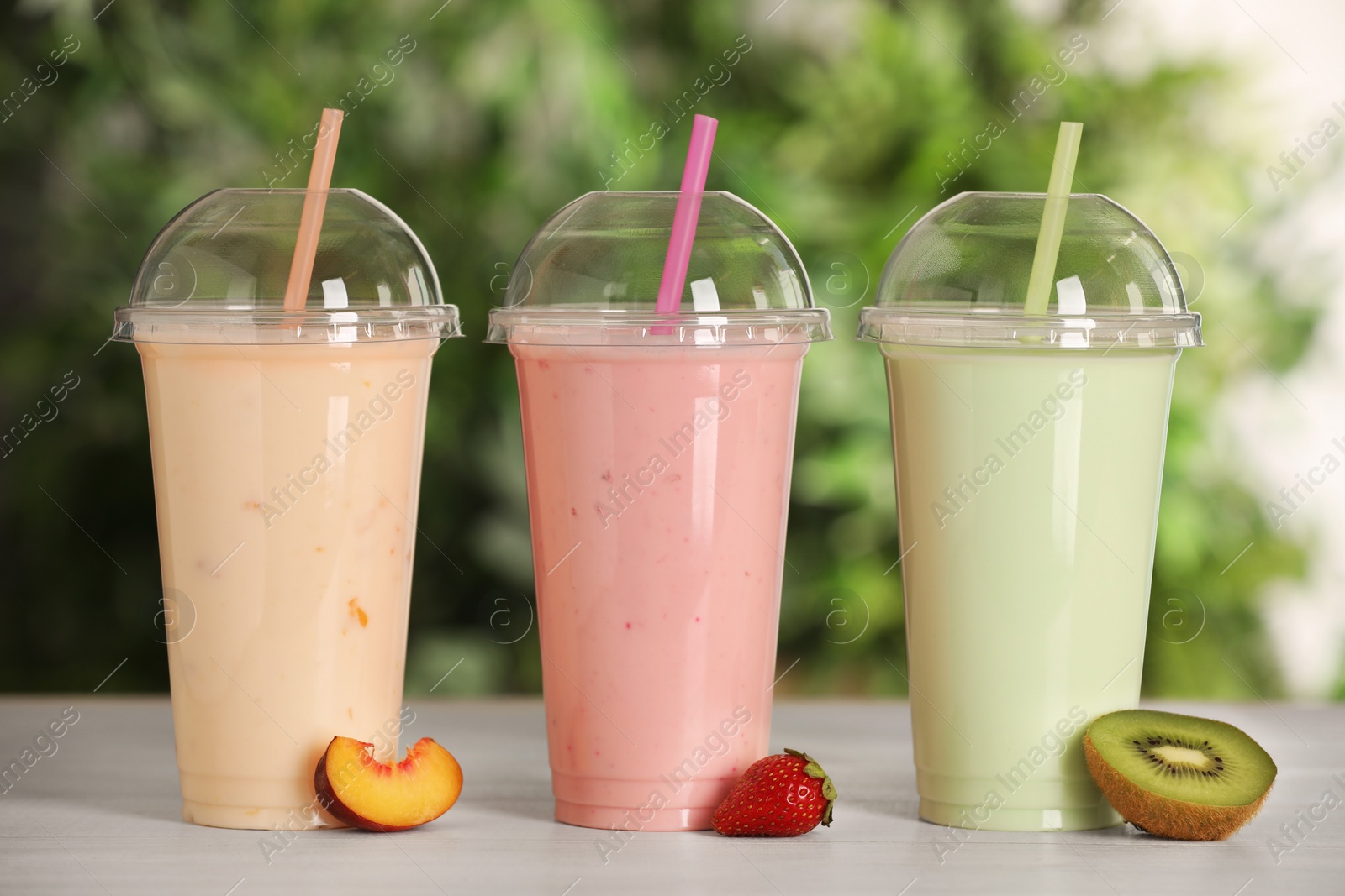 Photo of Plastic cups with different tasty smoothies and fresh fruits on wooden table outdoors