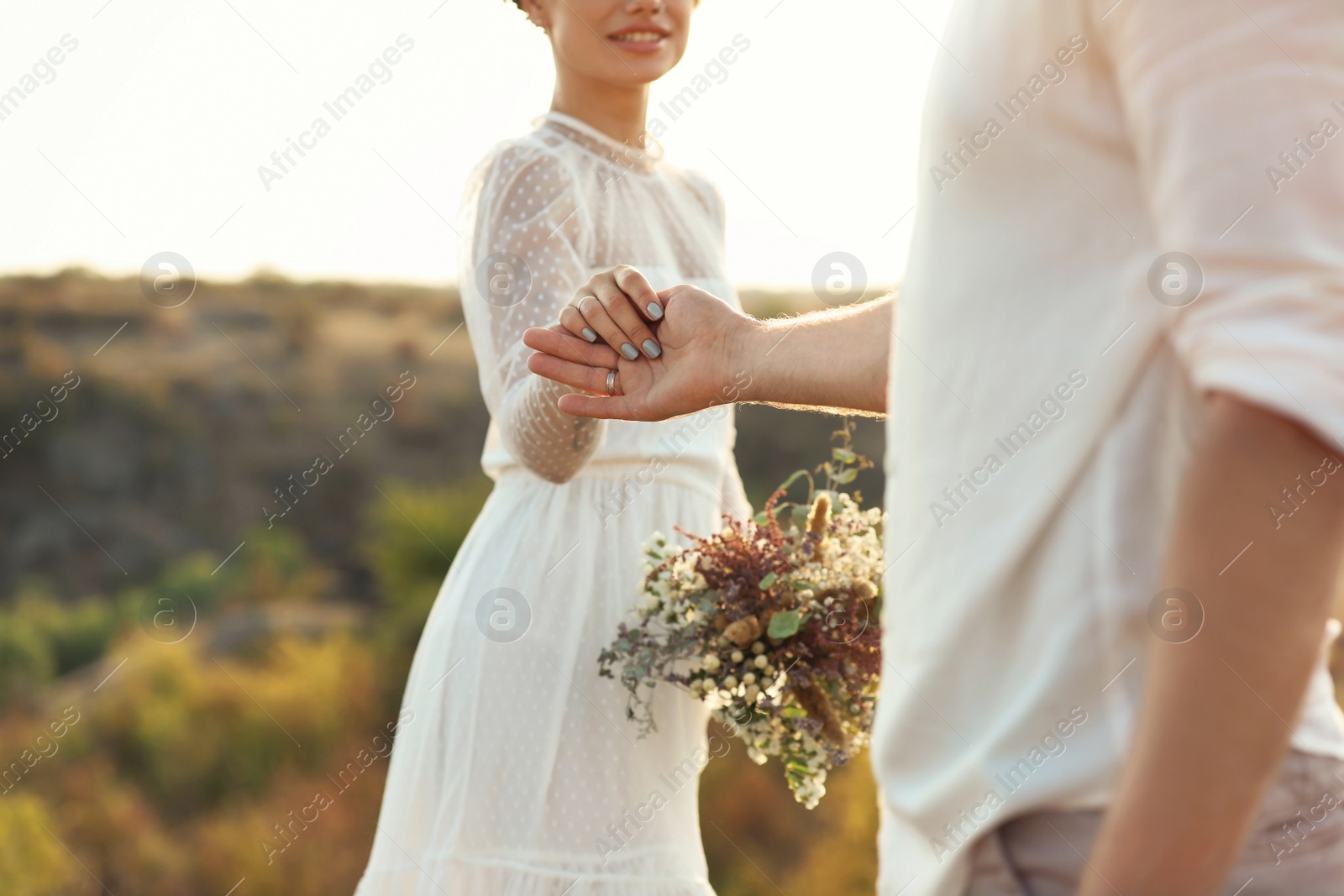 Photo of Happy newlyweds with beautiful field bouquet holding hands outdoors, closeup