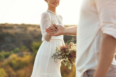 Photo of Happy newlyweds with beautiful field bouquet holding hands outdoors, closeup