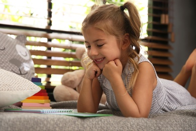 Cute little girl reading book near window at home