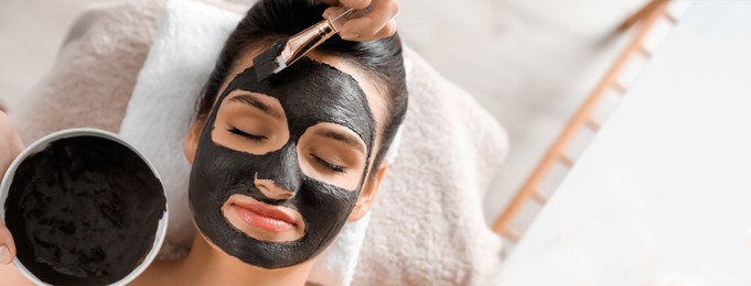 Cosmetologist applying black mask onto woman's face in spa salon, top view with space for text. Banner design