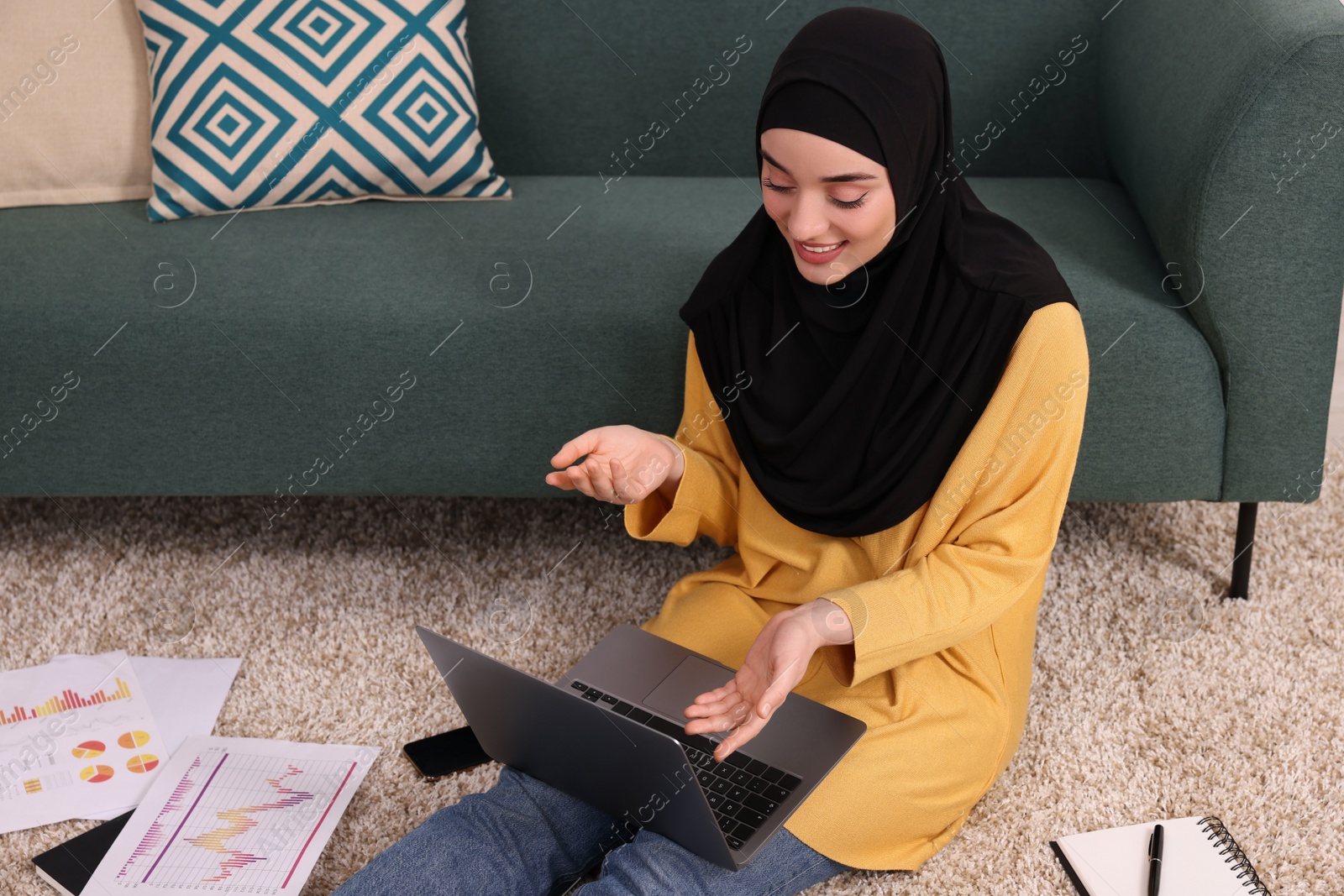 Photo of Muslim woman in hijab using video chat on laptop near sofa