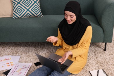 Muslim woman in hijab using video chat on laptop near sofa