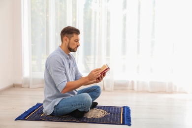 Photo of Muslim man reading Koran on prayer rug indoors