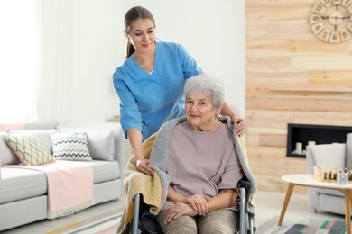 Photo of Nurse covering elderly woman in wheelchair with blanket indoors. Assisting senior people