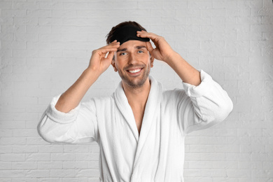 Photo of Happy young man in bathrobe and eye sleeping mask near white brick wall