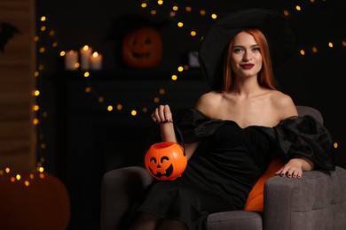 Photo of Young woman in scary witch costume with pumpkin bucket against blurred lights indoors, space for text. Halloween celebration