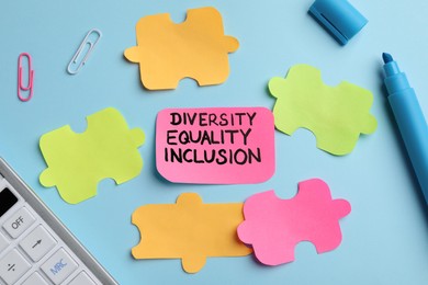 Photo of Sticky note with words Diversity, Equality, Inclusion and paper puzzle pieces on light blue background, flat lay