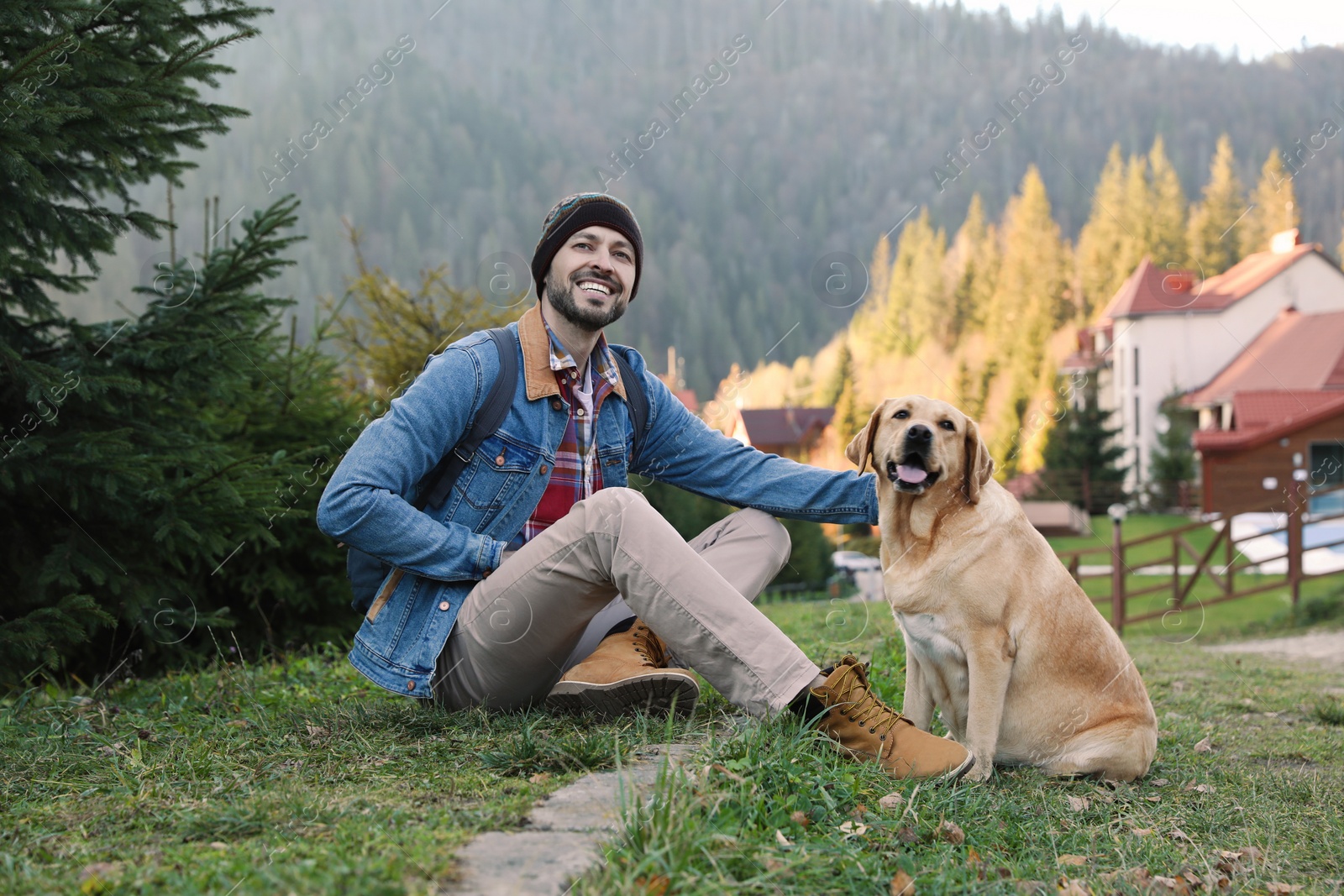 Photo of Happy man and adorable dog sitting on green grass in mountains. Traveling with pet