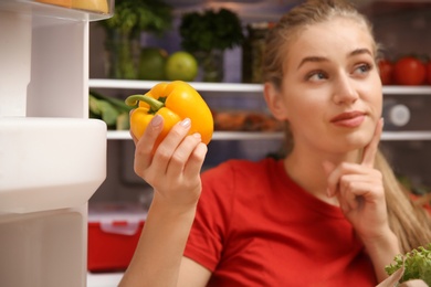 Photo of Young woman with fresh bell pepper near refrigerator at home