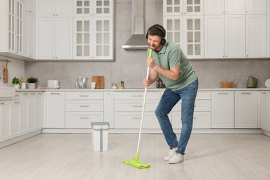 Photo of Enjoying cleaning. Happy man in headphones with mop singing while tidying up in kitchen