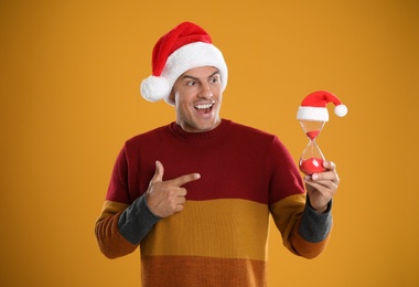 Photo of Man in Santa hat with hourglass on yellow background. Christmas countdown